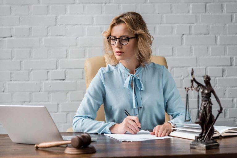 serious-female-judge-in-eyeglasses-working-with-papers-and-using-laptop-at-workplace